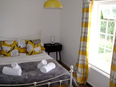 A bedroom with a bed with yellow and white floral pillows a bedside table with a lamp and a window with yellow and gray curtains at Phloxhaven in Reedham