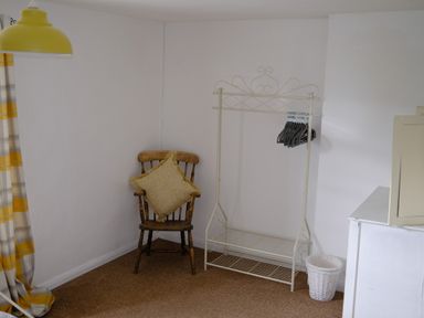 A corner of a room with a wooden chair holding a cushion a white metal clothing rack with hangers a white waste bin a white cabinet and yellow and gray curtains at Phloxhaven in Reedham