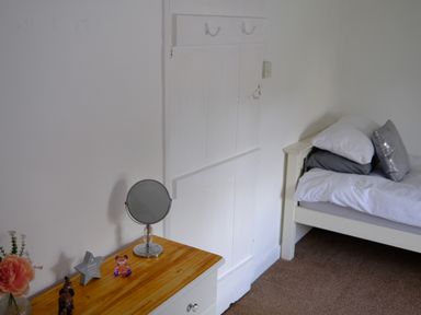 A bedroom corner with a white bed with pillows and a wooden dresser holding a round mirror and decorative items at Phloxhaven in Reedham
