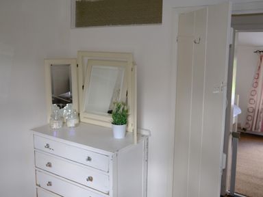 A white dresser with a three-paneled mirror jars and a potted plant on top next to a partially open door at Phloxhaven in Reedham