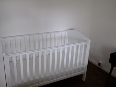A white baby crib with a mattress in a corner of a room with white walls and a carpeted floor at Phloxhaven in Reedham