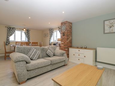 A living room with a gray sofa, wooden coffee table, white sideboard, brick chimney, dining table with chairs, and windows with patterned curtains at Bramley Cottage in Henley near Langport