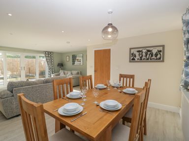 A dining table with six place settings and wooden chairs in a living room with grey sofas and a painting of cows at Bramley Cottage in Henley near Langport