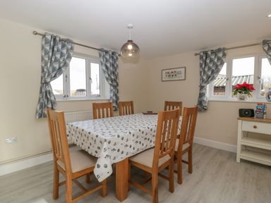 A dining room with a wooden table and six chairs with patterned curtains on windows at Bramley Cottage Henley near Langport