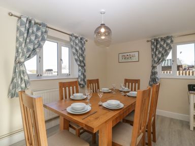 A dining room with a wooden table set for six near two windows with patterned curtains at Bramley Cottage in Henley near Langport