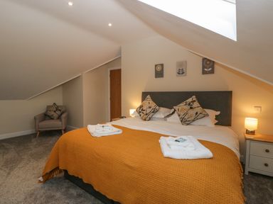A bedroom with a double bed covered with an orange blanket and two pillows two towels on bed a chair with a cushion a nightstand with a lamp and wooden door at Bramley Cottage Henley near Langport