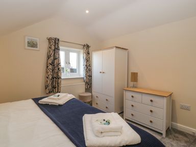 A bedroom with a bed with folded towels and toiletries a wardrobe a chest of drawers a stool and a window with patterned curtains at Bramley Cottage in Henley near Langport