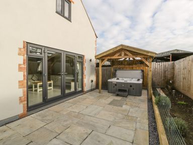 A patio area with a hot tub under a wooden pergola next to a house with glass doors and a garden bed at Bramley Cottage Henley near Langport
