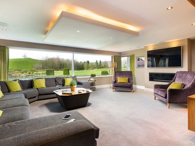 A living room with grey sofas, purple armchairs, a coffee table, a wall-mounted television, and large windows at Carus House at Carus Green Golf Club Kendal