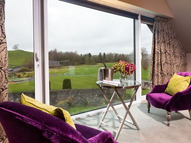 A seating area with two purple chairs and a small round table by a large window overlooking green fields at Carus House at Carus Green Golf Club in Kendal