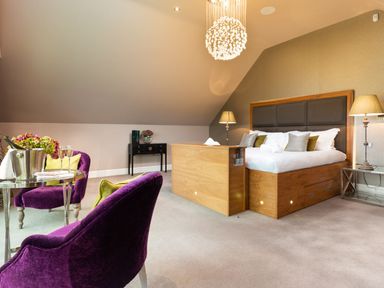 A bedroom with a wooden bed a chandelier purple chairs and a small table with champagne glasses at Carus House at Carus Green Golf Club in Kendal