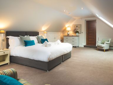 A bedroom with a large bed with white linens and pillows two bedside tables with lamps a chest of drawers an armchair and a wooden door at Carus House at Carus Green Golf Club Kendal