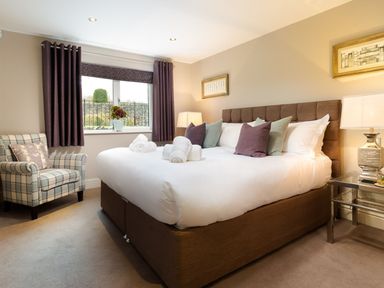 A bedroom with a large bed with white linens and pillows a plaid armchair and window with dark curtains at Carus House at Carus Green Golf Club Kendal