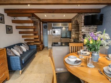 A dining area with a round wooden table set with dishes and a vase of flowers next to a kitchen with wooden beams and a blue sofa at Swallows Nest in Whitby