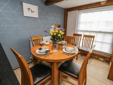 A dining area with a round wooden table set with plates bowls glasses and cutlery six wooden chairs a large window and a blue patterned wall at Swallows Nest in Whitby
