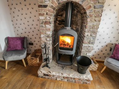 A wood burning stove set in a brick fireplace with two grey chairs and purple cushions on wooden floor at Swallows Nest in Whitby