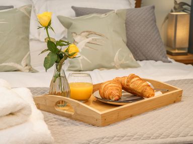 A wooden tray with two yellow roses in a glass vase a glass of orange juice and two croissants on a bed with pillows at Swallows Nest in Whitby