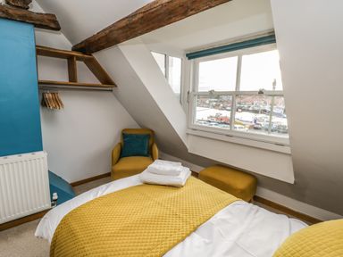 A bedroom with a bed covered in yellow bedding next to a window with a harbour view a chair and footstool in the corner at Swallows Nest in Whitby