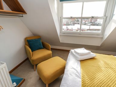 A bedroom corner with a yellow armchair and ottoman a bed with folded white towels and a window showing a harbor view at Swallows Nest in Whitby