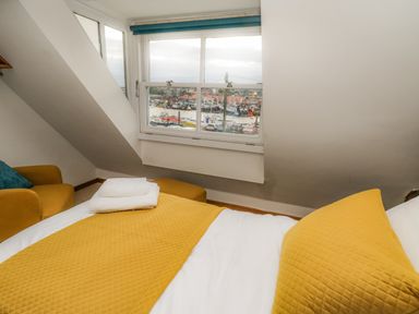 A bedroom with a bed covered by a yellow quilt and pillow towels on the bed a yellow armchair near a window showing a view of a harbor at Swallows Nest Whitby