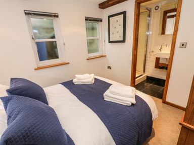 A bedroom with a double bed covered with blue pillows and blanket two windows a framed picture on the wall and a door leading to a bathroom at Swallows Nest Whitby