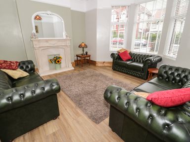 A living room with three dark green leather sofas a beige rug wooden flooring a white fireplace with a mirror and a bouquet of sunflowers on it a side table with a lamp and large windows with white blinds at 23 Carmen Sylva Road in Llandudno