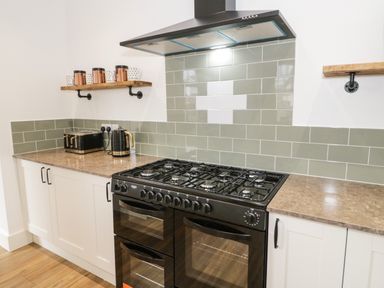 A kitchen with a black stove and oven white cabinets green tiled backsplash and wooden shelves with mugs and containers at 23 Carmen Sylva Road in Llandudno