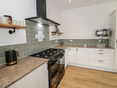A kitchen with a black stove, white cabinets, wooden countertops, green tile backsplash, a kettle, mugs, and a microwave at 23 Carmen Sylva Road in Llandudno