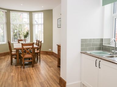 A dining area with a wooden table and six chairs next to windows with white blinds and a kitchen sink with cabinets and green tiled backsplash at 23 Carmen Sylva Road in Llandudno