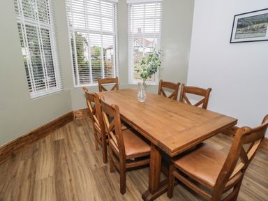 A dining area with a wooden table and six chairs near large windows with blinds at 23 Carmen Sylva Road in Llandudno