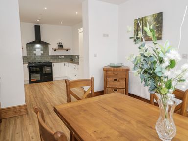 A dining area with a wooden table and chairs next to a kitchen with white cabinets and a black stove at 23 Carmen Sylva Road in Llandudno