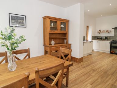 A dining area with a wooden table and chairs a vase with flowers a wooden cabinet with glass doors and a kitchen with white cabinets and appliances at 23 Carmen Sylva Road in Llandudno