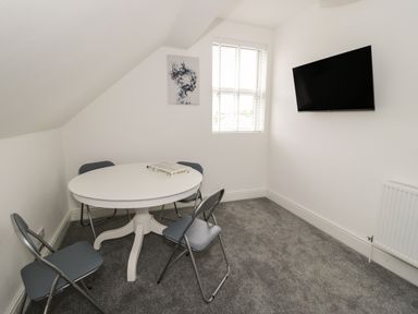 A room with a round white table and four gray folding chairs a window with blinds and a wall-mounted TV at 23 Carmen Sylva Road in Llandudno