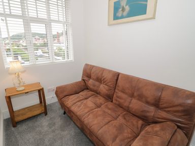 A small room with a brown leather sofa a wooden side table with a lamp and a window with white blinds at 23 Carmen Sylva Road in Llandudno