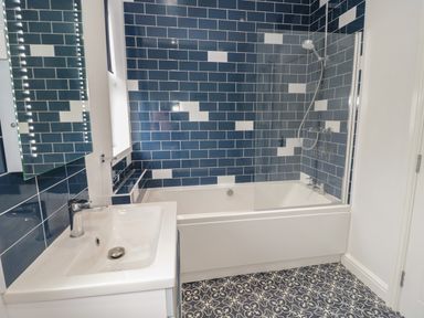 A bathroom with blue and white tiled walls a white bathtub with shower and sink at 23 Carmen Sylva Road in Llandudno
