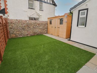 A small backyard with artificial grass a stone wall a wooden shed and a paved path at 23 Carmen Sylva Road in Llandudno
