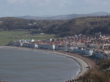 A coastal town with a curved beachfront promenade lined with buildings and hills in the background at 23 Carmen Sylva Road in Llandudno