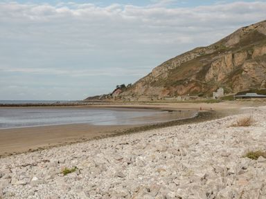 A rocky shoreline leading to a sandy beach with cliffs and buildings in the background at 23 Carmen Sylva Road in Llandudno