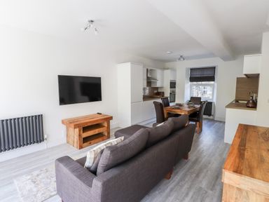An open plan living room and kitchen with a gray sofa facing a wall mounted TV wooden dining table with four chairs and white kitchen cabinets at 8 Waterhead Terrace Ambleside