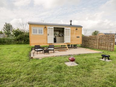 A small wooden cabin with open double doors on a gravel patio with two chairs a table and a red grill on grass at Waylands in Uffington