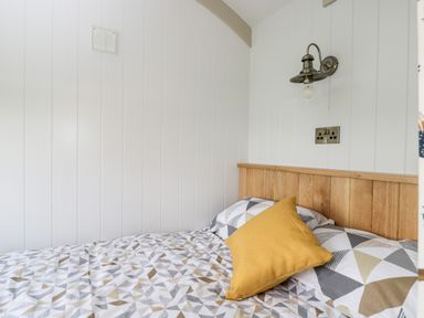 A bed with patterned bedding and a yellow pillow against a white paneled wall with a wall lamp and electrical outlet at Waylands in Uffington