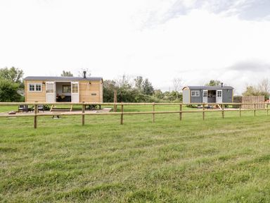 Two small wooden cabins on wheels with open doors and outdoor seating areas behind a wooden fence in a grassy field at Waylands in Uffington
