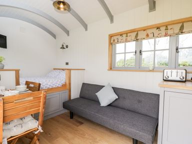 A room with a wooden bed platform with bedding a gray sofa with a pillow a wooden dining table and chairs a window with a patterned blind and a toaster on a counter at Ridgeway in Uffington