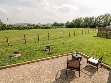 A gravel patio area with two brown chairs and a small table with wine and two glasses next to a grassy yard with a wooden fence and open field at Ridgeway in Uffington