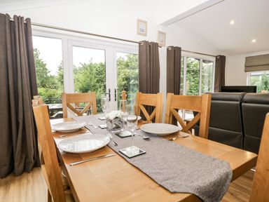 A dining table with plates glasses and cutlery in a room with large windows and brown curtains at Robin Lodge in Cartmel