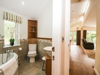A bathroom with a bathtub toilet and sink adjacent to a living room with blue armchairs and brown curtains at Robin Lodge in Cartmel