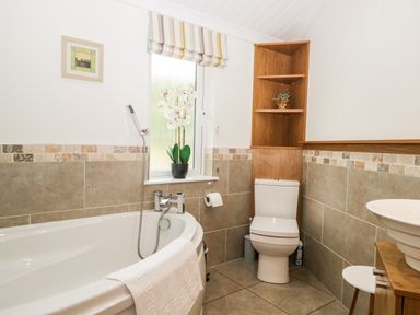 A bathroom with a bathtub a toilet and a sink with wooden shelves and tiled walls at Robin Lodge in Cartmel