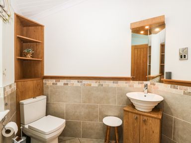 A bathroom with a toilet, a wooden cabinet with a white sink, a stool, and a mirror at Robin Lodge in Cartmel