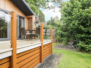 A wooden deck with glass railing and outdoor furniture next to a house with a garden and green trees at Robin Lodge in Cartmel