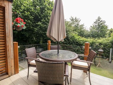 An outdoor deck with a round glass table and four wicker chairs with cushions at Robin Lodge in Cartmel